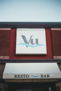 Vu restaurant sign on brick exterior featuring a white square with a gray 'Vu' and a blue wave motif, above a white canopy that reads RESTO BAR
