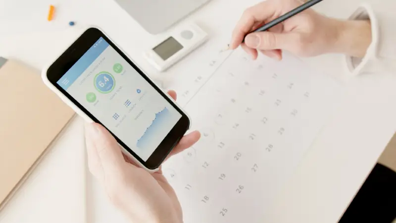 Small Business Marketing Calendar » Sparks Signs and Graphics Person holds a smartphone showing a health app with a 6.4 score, while a hand writes on a calendar with a pencil in the background.