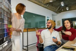 Three colleagues in a bright design studio review a wall of color swatches; a standing woman points to samples while two seated coworkers consult with a card