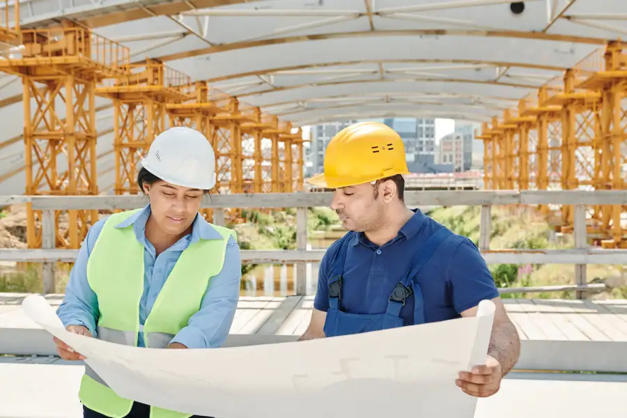 Two construction workers review a large blueprint on a sunny site with orange steel framework in the background.