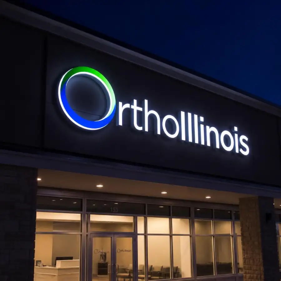 OrthoIllinois clinic storefront at night with a glowing circular blue-green logo and large illuminated sign on the front.