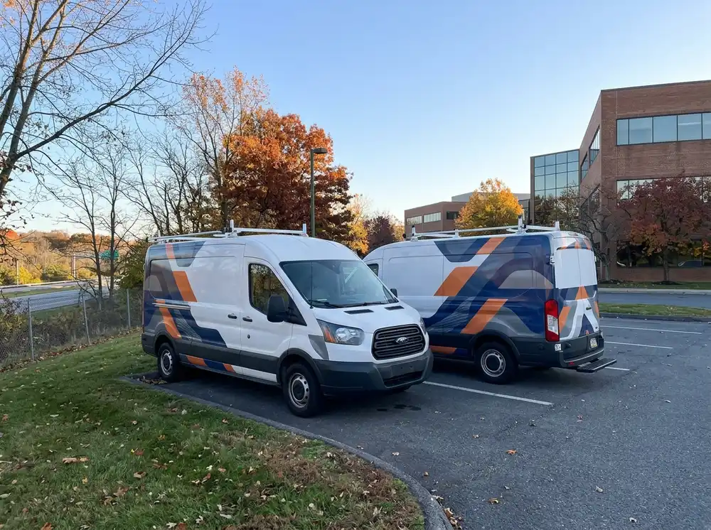 Two white vans with blue and orange diagonal graphics parked in a lot beside a brick office building amid autumn trees.