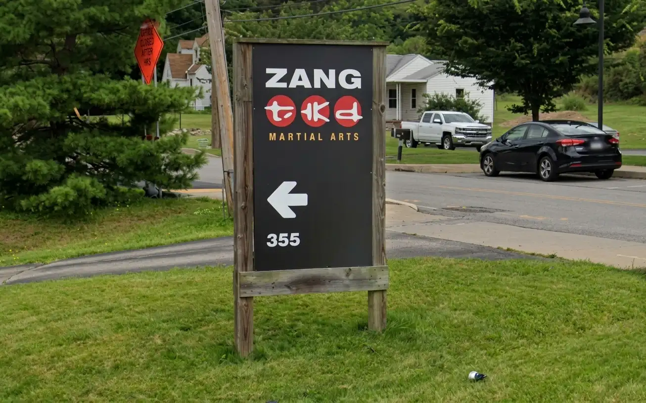 Outdoor black post-frame directional sign for Zang TKD Martial Arts with white logo, arrow, and address number installed roadside, by Spark Signs in Warrendale PA