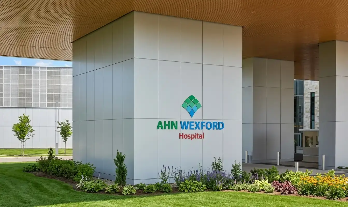 Modern hospital exterior with AHN Wexford Hospital sign on a gray pillar and landscaped garden in the foreground, sunny day